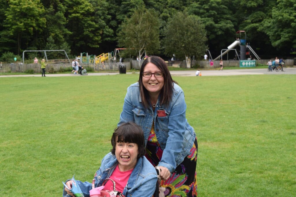 Two ladies smile for a photo in a park. The lady in the front is in a wheelchair. Both are wearing denim jackets.