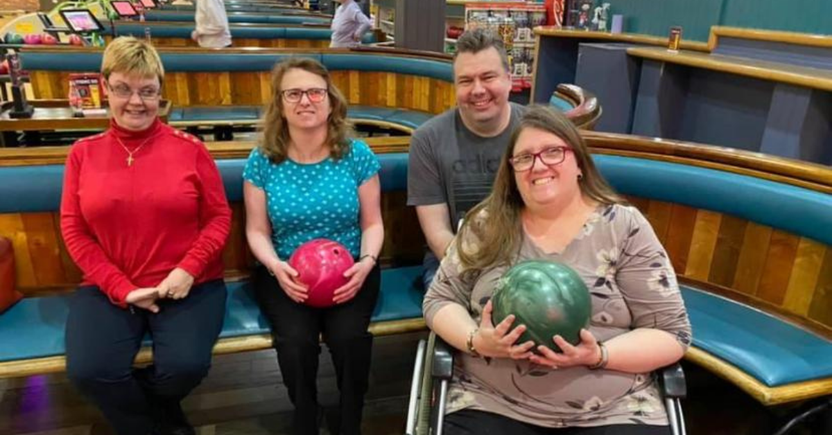Four adults pose for a photo at a bowling alley. One is a wheelchair user and they are holding a green bowling ball.