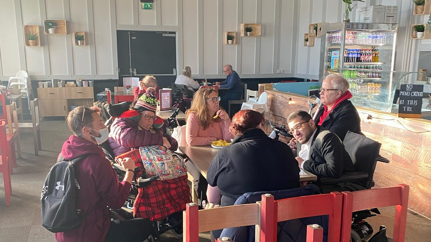A group of SHASBAH members, who have various disabilities, gather around a table in a cafe to eat and socialise together.
