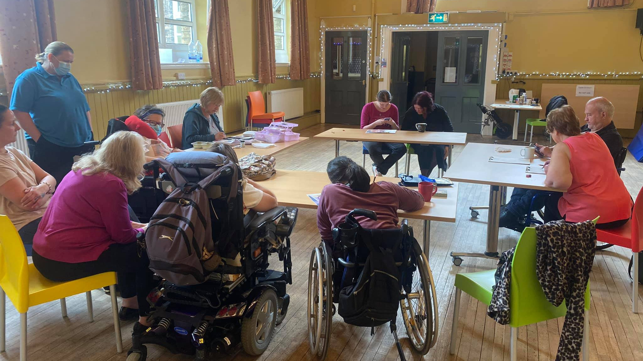 A group of people, some of whom are wheelchair users, sit around a circle of tables and work on their own projects.