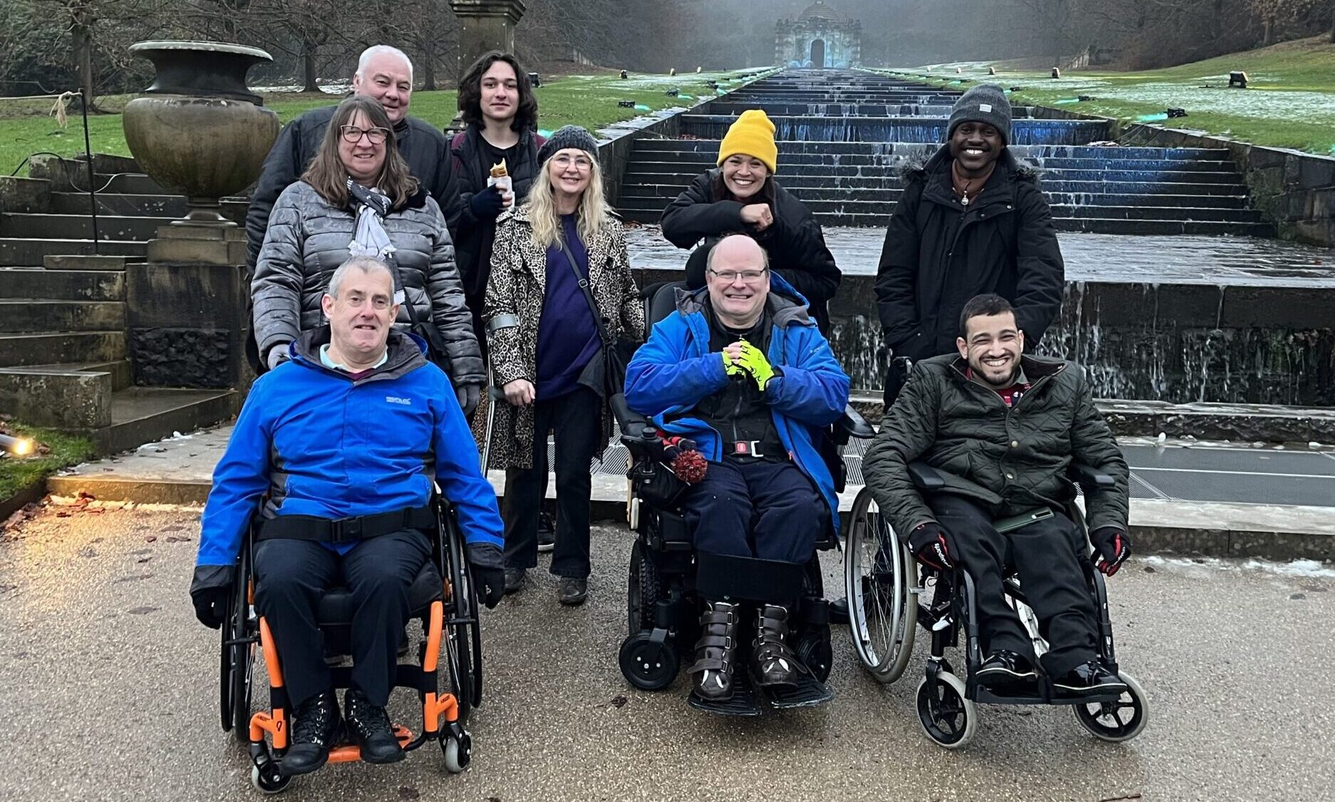 A group of charity members, friends and volunteers smile for a photo in front of a fountain. In the front row are three men who are using wheelchairs and have spina bifida.