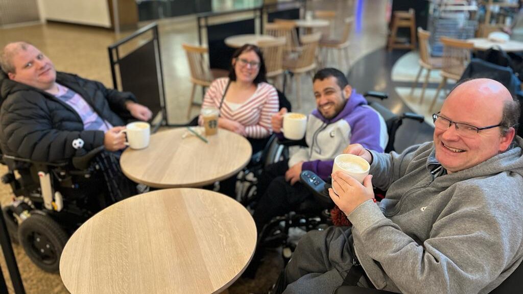 A group of SHASBAH members smile for a photo while drinking hot drinks in an accessible cafe.