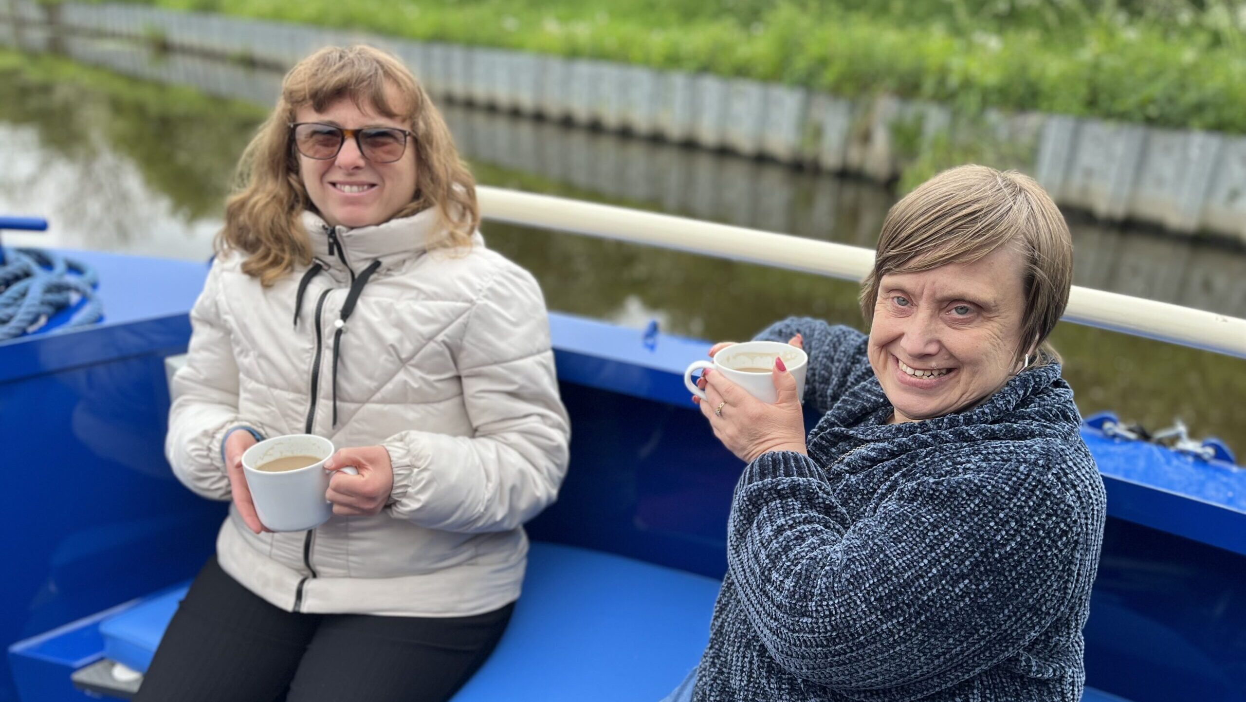 Two women are smiling for the camera. Each one is holding a cup of tea and they are on an accessible canal boat trip.